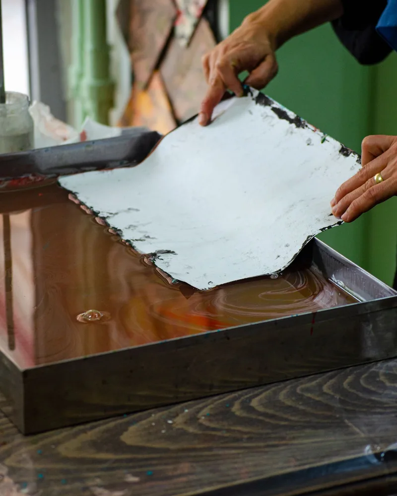 Manos levantando una hoja de papel húmeda sobre una cubeta poco profunda en una mesa de taller.